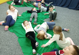 Dzieci w różnych pozycjach leżą na podłodze na zielonej tkaninie.
